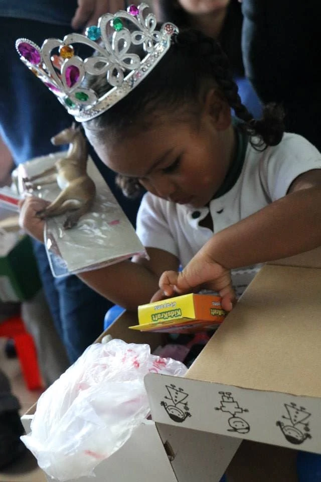 Hispanic girl wearing a princess crown opens a shoebox fill of toys. 