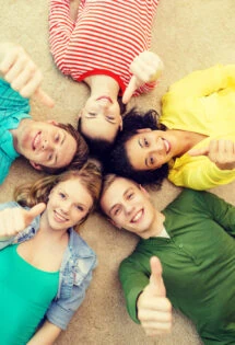 Group of young friends lying on the floor with heads together, smiling and giving a thumbs up.
