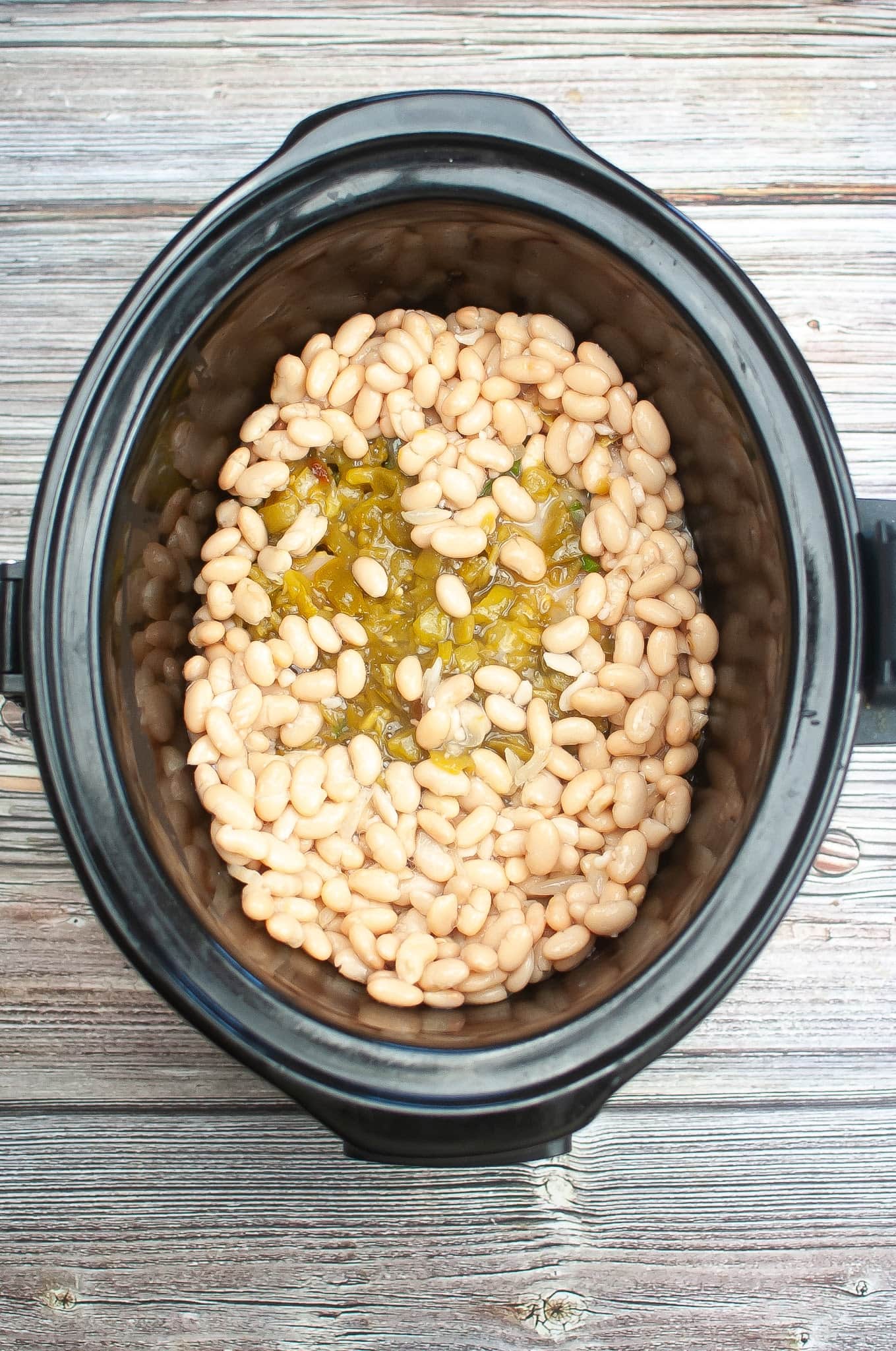 A black crock pot with white beans and green peppers for a white chicken chili. 