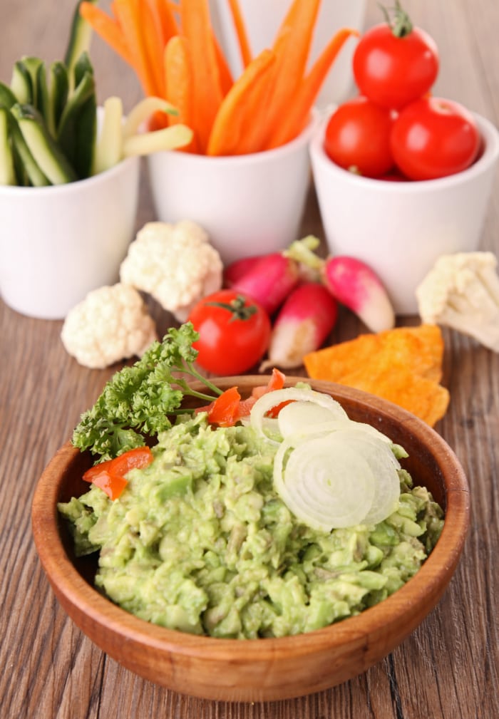A bowl of guacamole is in the forefront of the picture with cucumber spears, carrot spears, cherry tomatoes, cauliflower florets, and radishes behind the wooden bowl.