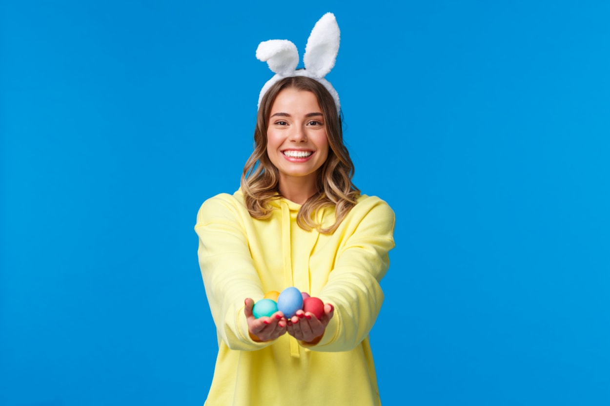 A young woman wearing bunny ears and holding Easter eggs on a blue background to illustrate Easter basket ideas for college students. 