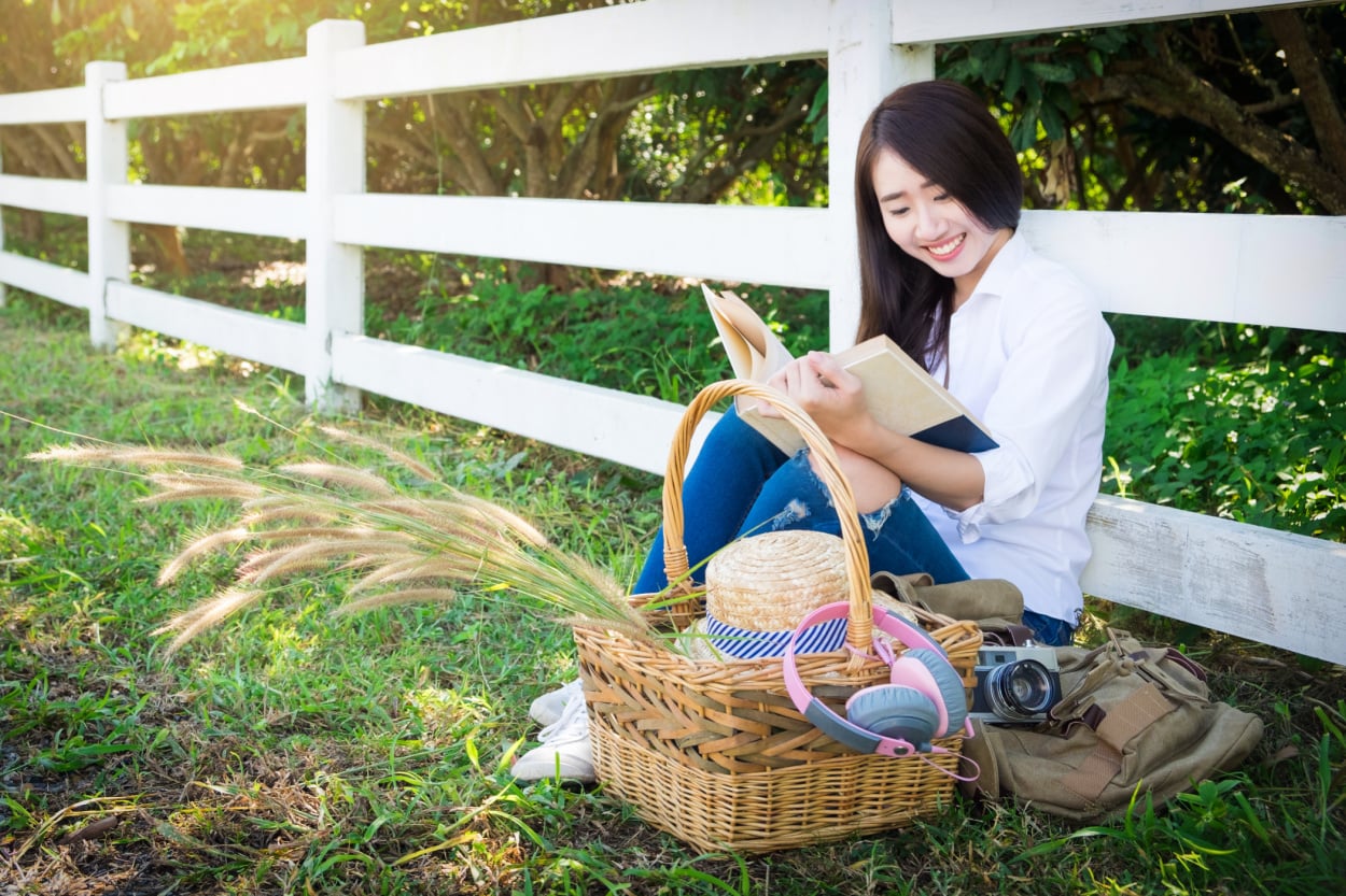 A young asian teenage girl sitting on the grass reading a book with an Easter basket beside her.