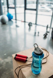 A water bottle and jump rope on a wooden surface in a gym with exercise equipment in the background.