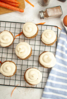 Freshly frosted grain-free carrot cake cupcakes on a cooling rack with ingredients and a striped towel in the background.