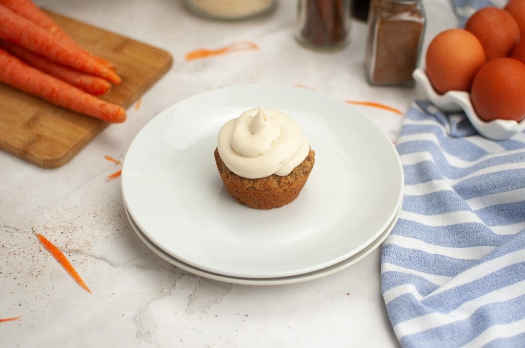 A grain-free carrot cupcake topped with cream cheese frosting on a white plate, with ingredients in the background.