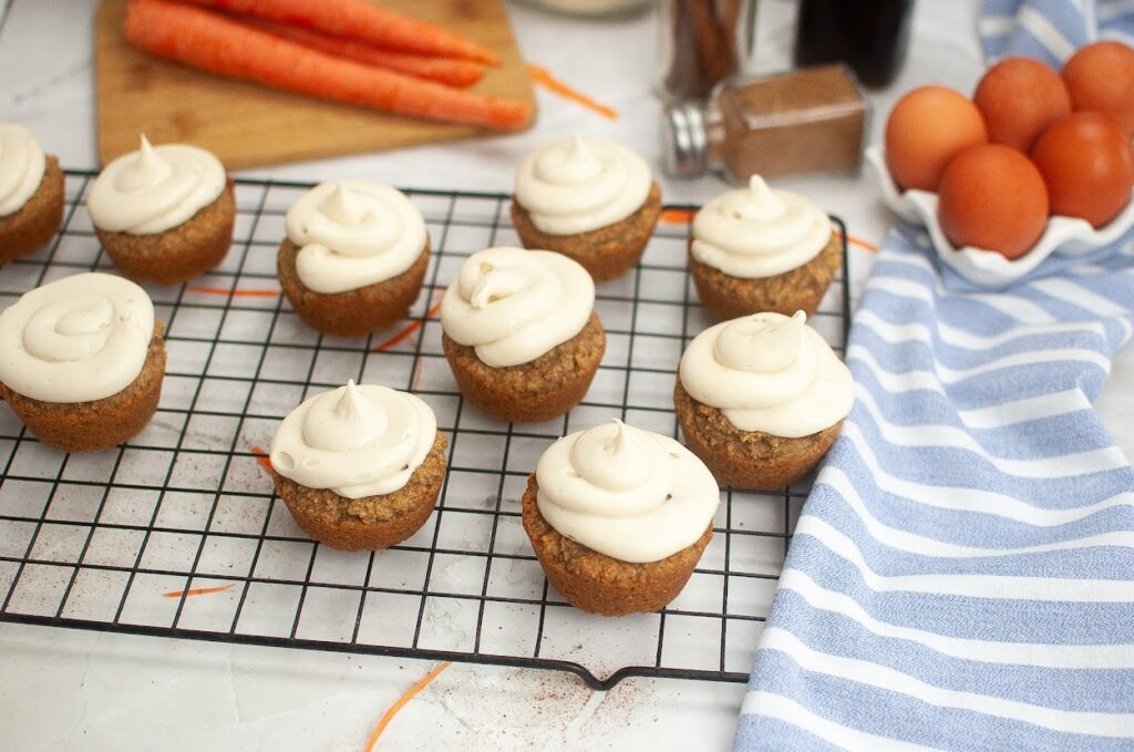 Grain-free carrot cupcakes with cream cheese frosting on a wire rack, ingredients nearby on a kitchen counter.