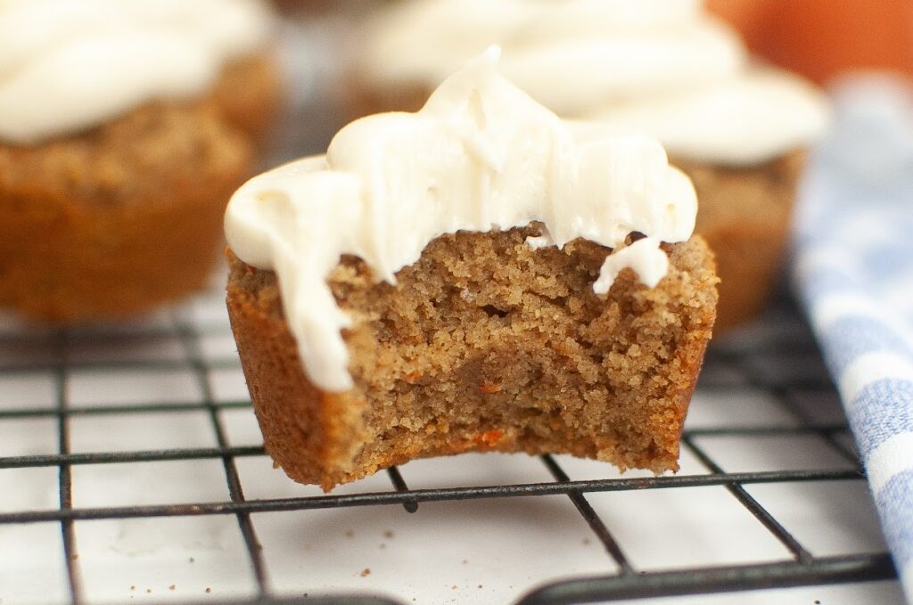 A freshly baked frosted grain-free carrot cake muffin with a bite taken out, displayed on a cooling rack.