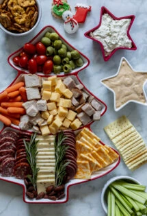 A festive holiday spread featuring an assortment of cheeses, charcuterie, crackers, and vegetables arranged in star and tree-shaped dishes.