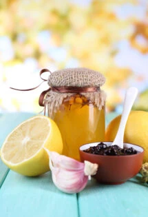 A jar of honey with a spoon, flanked by a cut lemon, garlic, and a small bowl of raisins on a wooden table.