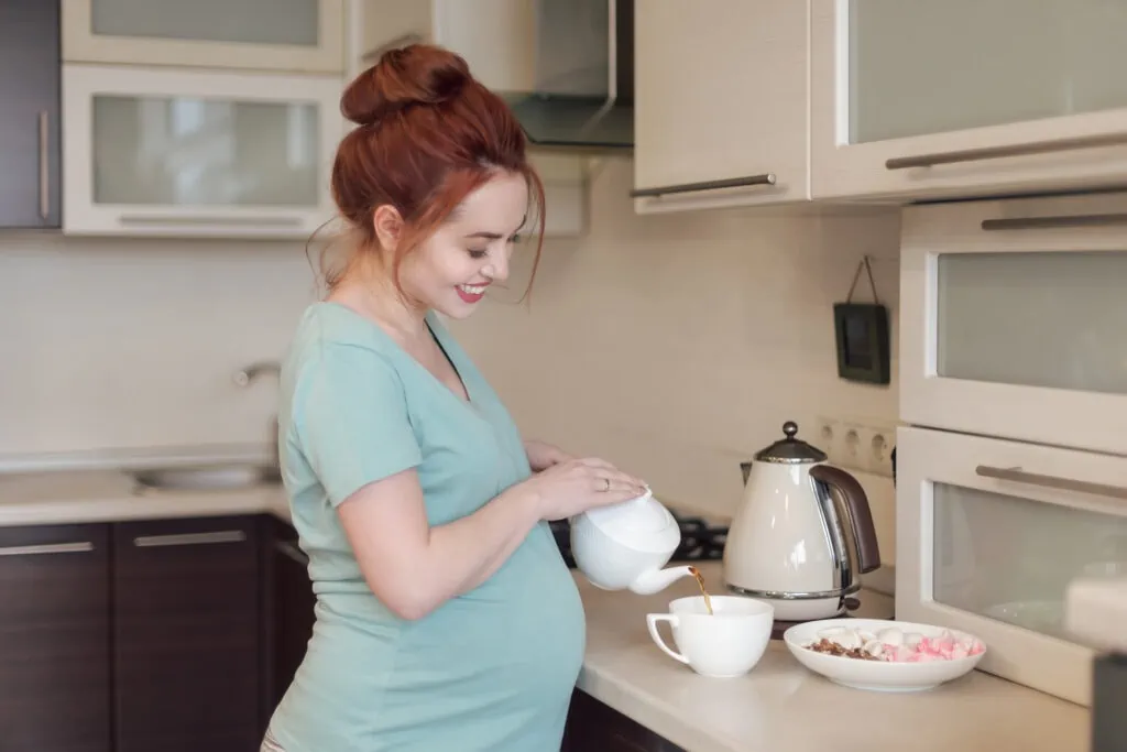 a pregnant redheaded woman pouring herself a cup of tea in her uncluttered kitchen