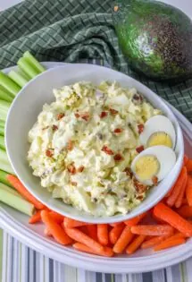 A bowl of egg salad with avocado and bacon, accompanied by celery sticks, baby carrots, and an avocado.