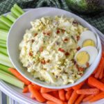 A bowl of egg salad garnished with egg and bacon, accompanied by celery sticks and baby carrots.