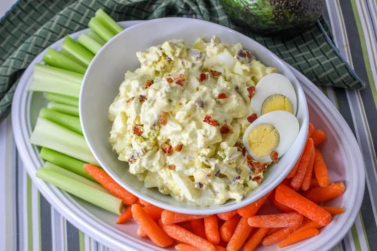 A bowl of egg salad garnished with egg and bacon, accompanied by celery sticks and baby carrots.