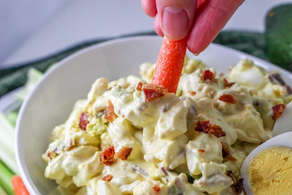 A person dipping a carrot stick into a bowl of creamy salad with bacon bits.