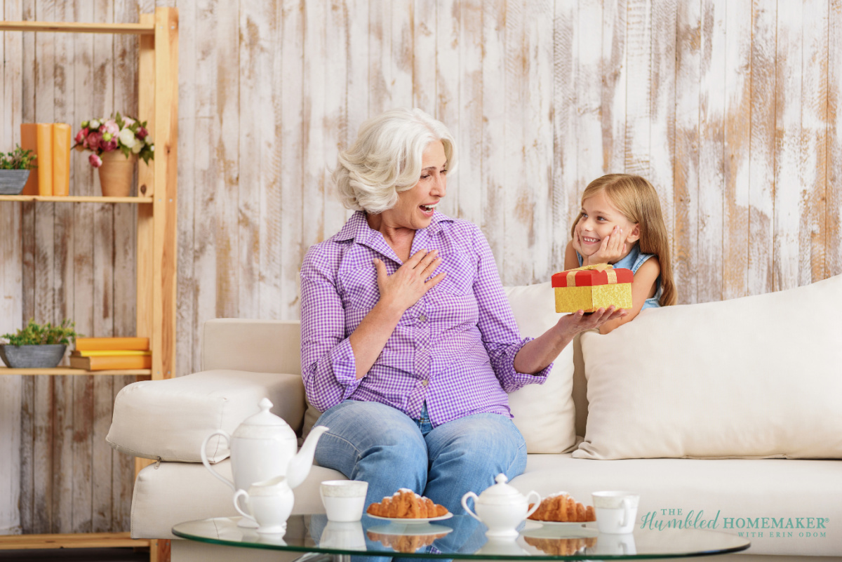 A grandmother receiving a mother's day gift from her young granddaughter, sitting together on a couch with snacks on the coffee table in a cozy room.