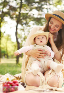 A mother in a sunhat smiles at her baby on a picnic blanket in a sunny park, surrounded by wildflowers and a Mother's Day basket of fruit.
