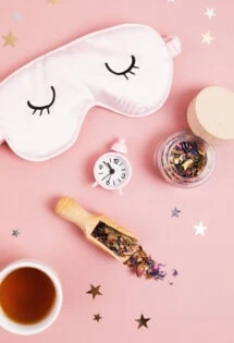Flat lay composition with a sleep mask, alarm clock, a cup of tea, a jar of herbal mix, and a wooden scoop on a pink background with star confetti.