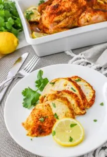 Roasted chicken slices served on a white plate garnished with lemon and parsley, with more chicken in the background.