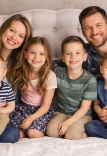 A family of two adults and four children sit together on a bed, all smiling at the camera.