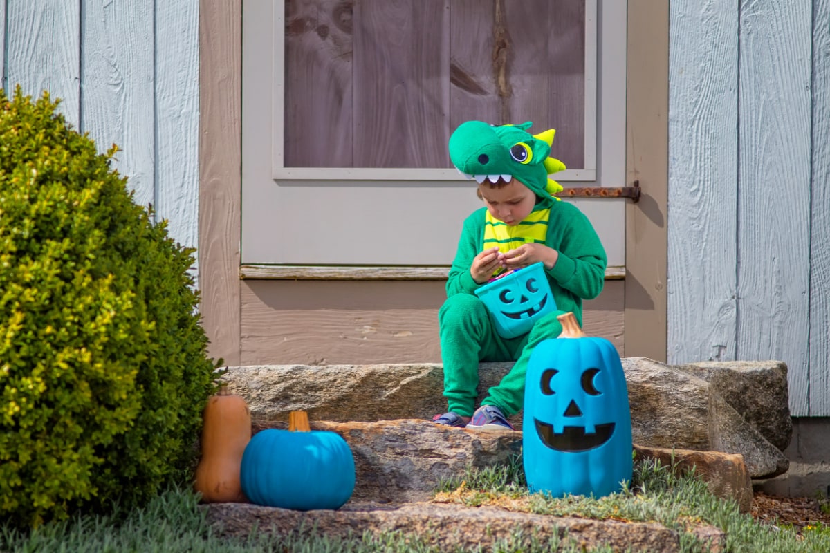 A child in a blue dinosaur costume sits on the steps of a house, excitedly awaiting non-food Halloween treats for a post on candy alternatives for Halloween.