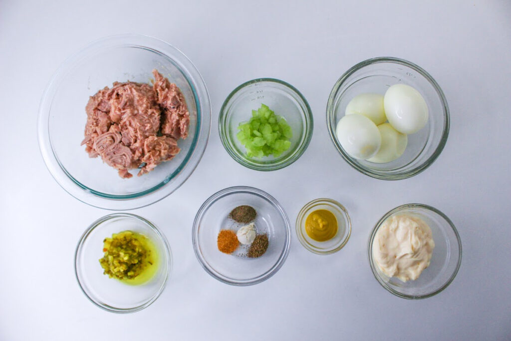 Ingredients for making tuna salad laid out in separate prep bowls on a white surface.