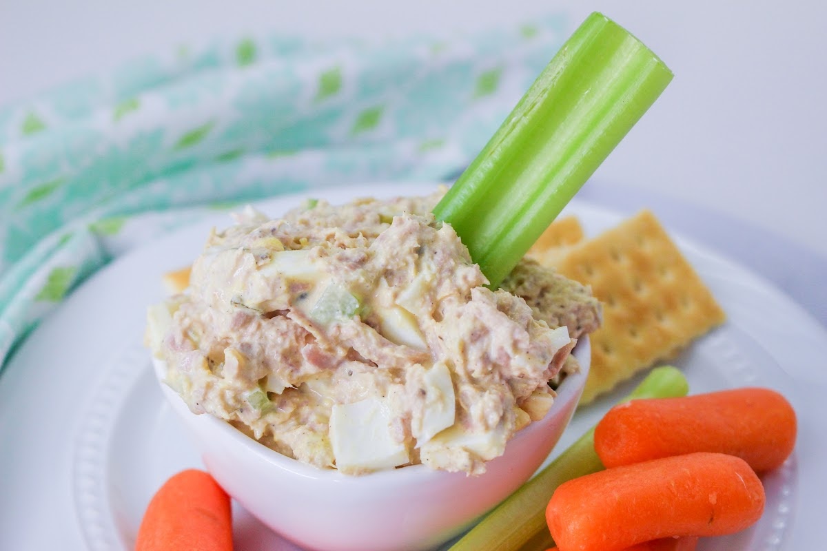 A bowl of low carb tuna salad garnished with a celery stick, surrounded by baby carrots and crackers on a white plate.