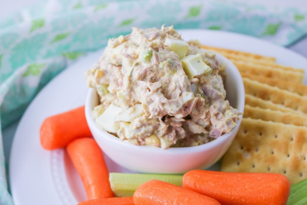 A bowl of tuna salad served with crackers and fresh carrot and celery sticks.