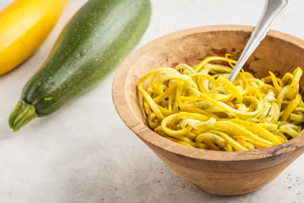 a wooden bowl with zucchini noodles inside of it along with a fork since zucchini noodles, also called zoodles, are a great, low-carb pasta alternative. There is also a picture of an uncut green zucchini and an uncut yellow squash in the background. 