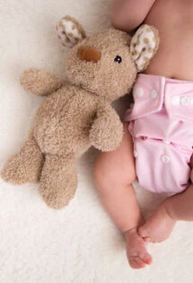 Baby in a pink cloth diaper lying next to a plush bear toy.