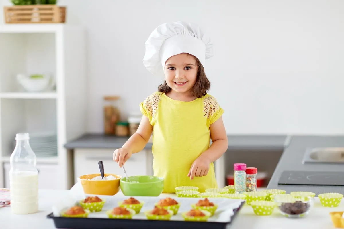 A little girl in a chef's hat baking cupcakes in the kitchen, exploring non-toy gift ideas.