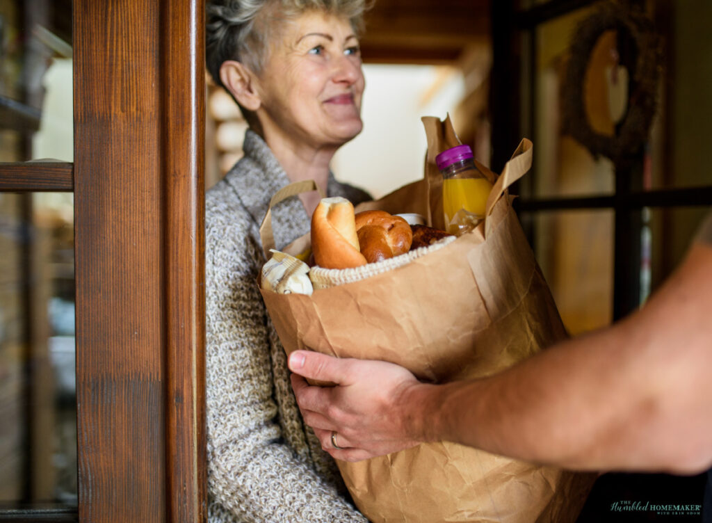 A smiling elderly woman receives a grocery delivery from a person at the door.