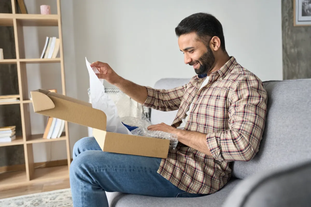 A man sits on a couch with a subscription box in front of him. This box looks similar to the boxes sent from Stitch Fix for men. 