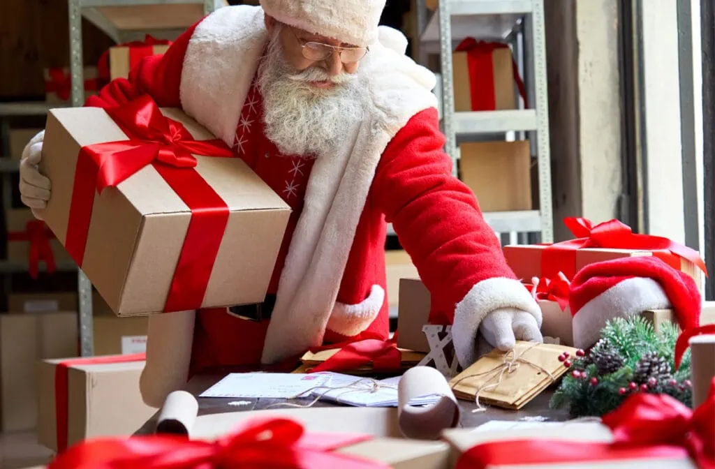 Santa Claus is surrounded by boxes of gifts in a warehouse.