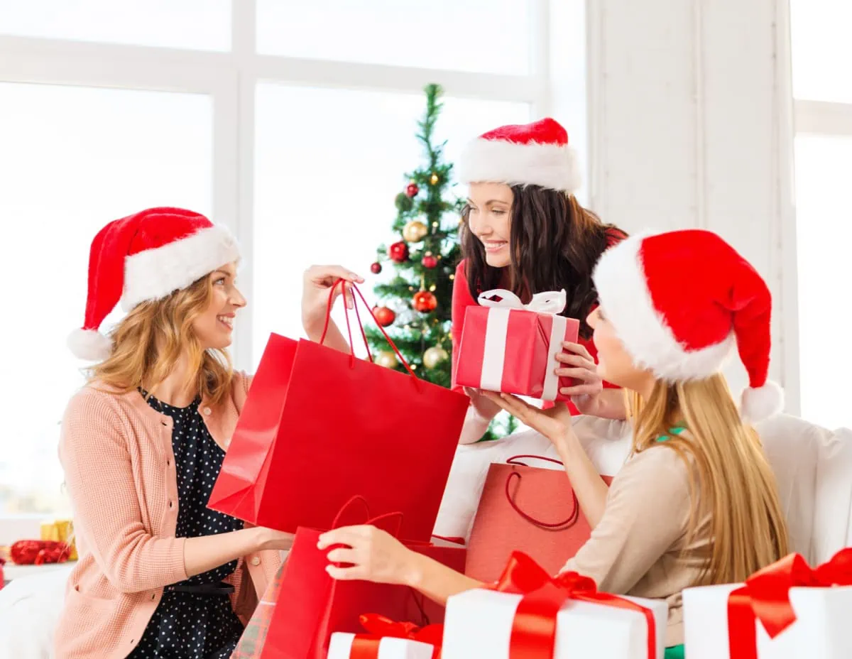 Three women in santa hats engaging in simple gift-giving, holding festive gift bags.