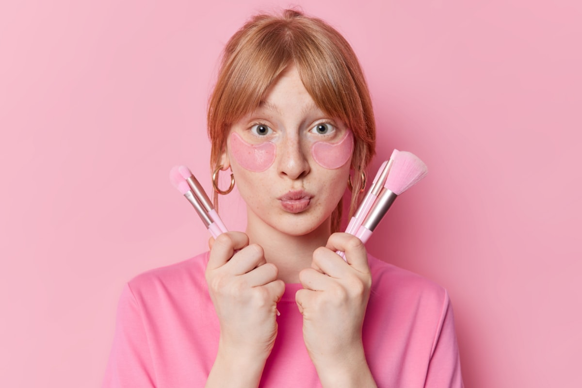 A young girl with pink makeup brushes on a pink background.