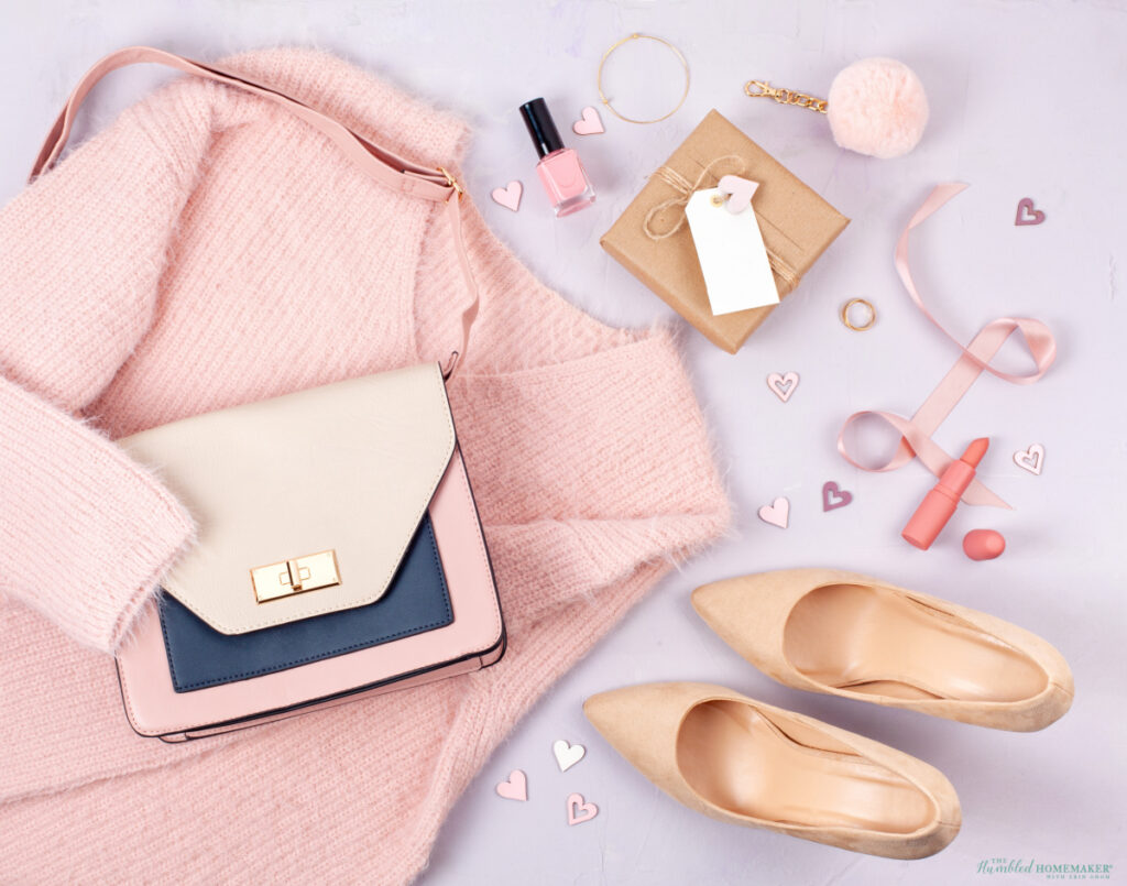 Flat lay of a stylish ensemble featuring a pink sweater, pastel handbag, ballet flats, makeup items, and decorative hearts